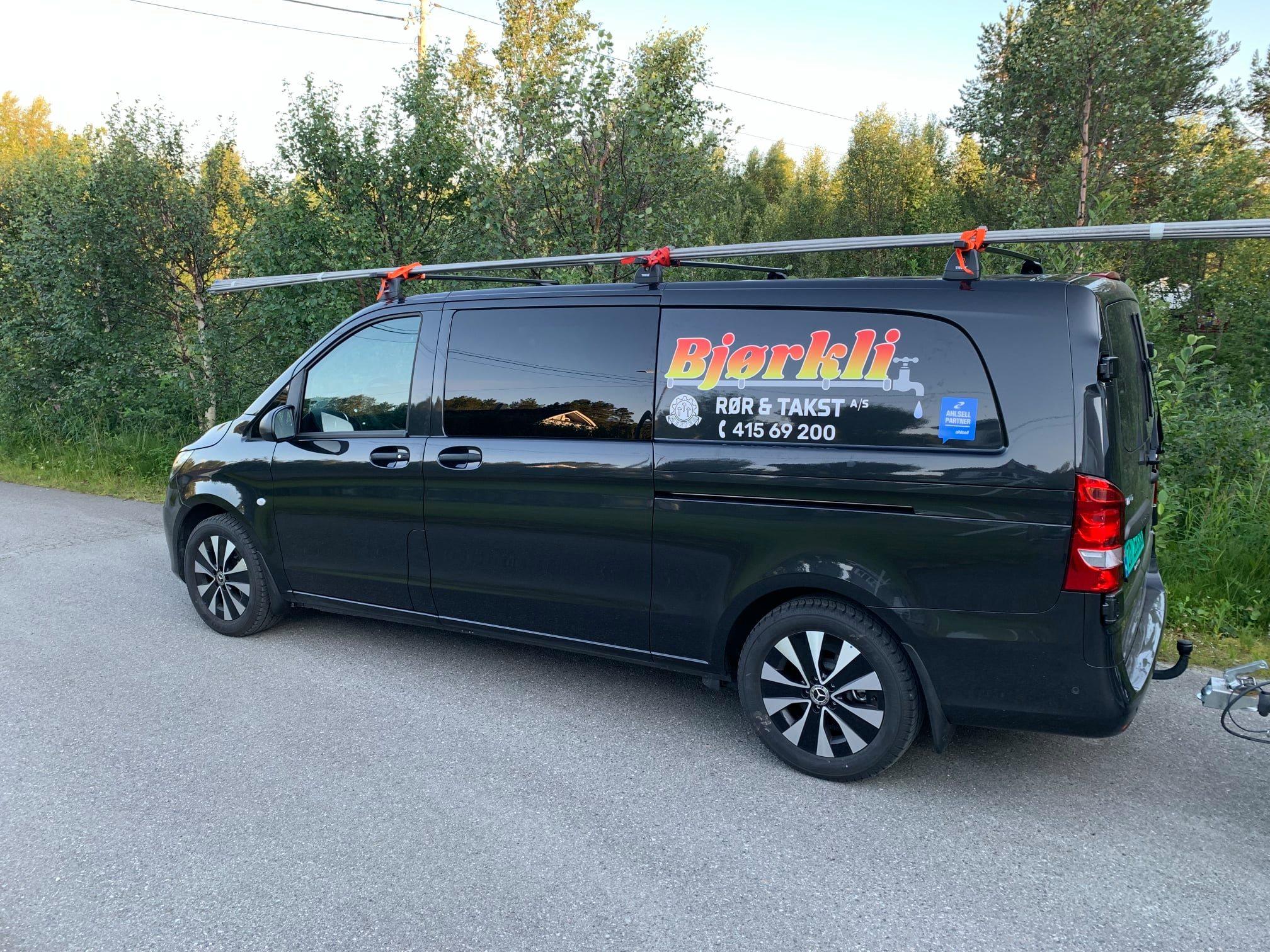 En svart varebil med takstativ og logoen "Bjørklit" på siden, parkert langs en vei i naturen.