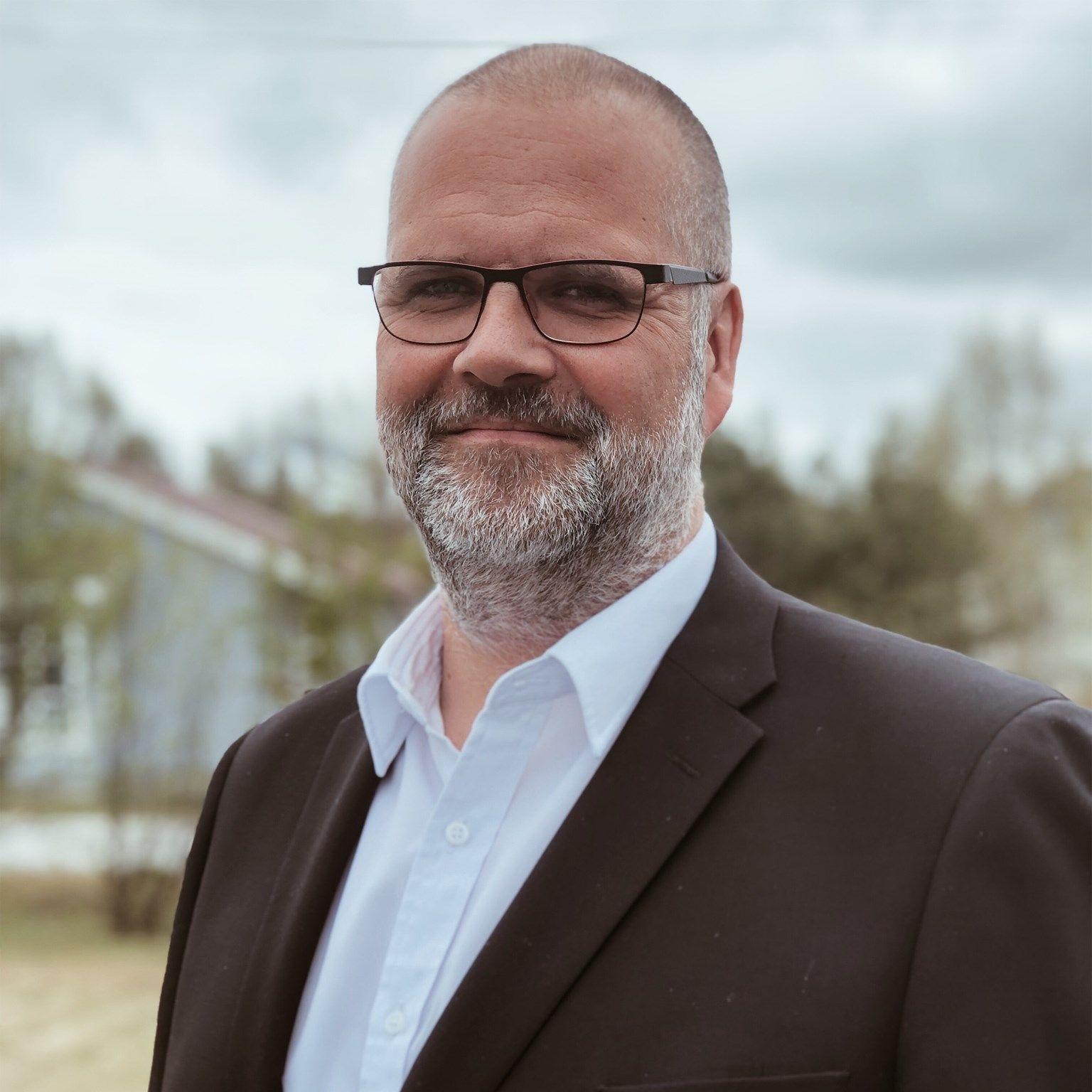 En mann med skjegg og briller iført dress står ute med trær og hus i bakgrunnen.