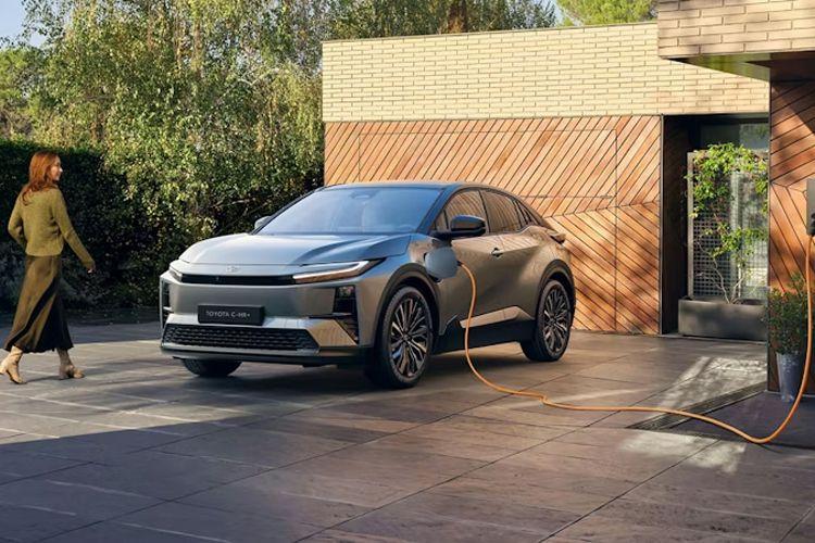 En moderne elbil parkert foran et hus med en kvinne som går forbi. Ladeledning er koblet til bilen.