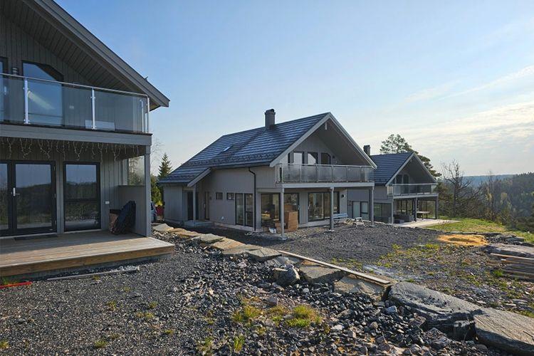 Moderne hus med store vinduer og balkonger, omgitt av natur og steinlagt område. Solfylt himmel i bakgrunnen.