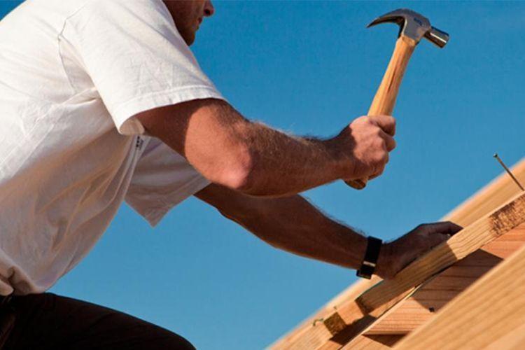 En håndverker som bruker en hammer til å feste treverk på taket under en klar blå himmel.