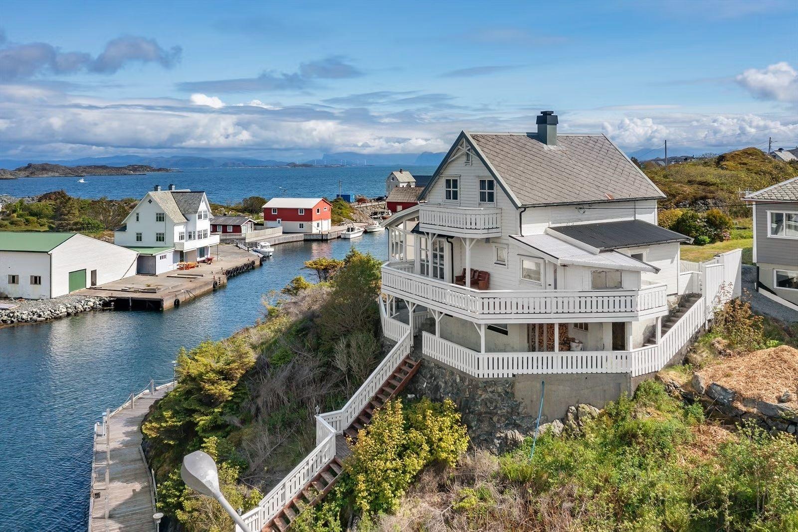 En vakker utsikt over et hvitt hus med terrasse ved en havn, omgitt av fjell og småbåter.