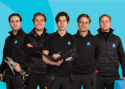 Fem håndverkere i uniform med verktøy og notatblokk, smilende foran en blå bakgrunn.