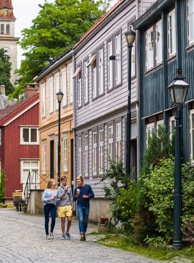 Tre personer går langs en brosteinsbelagt gate med fargerike trehus og gatebelysning.