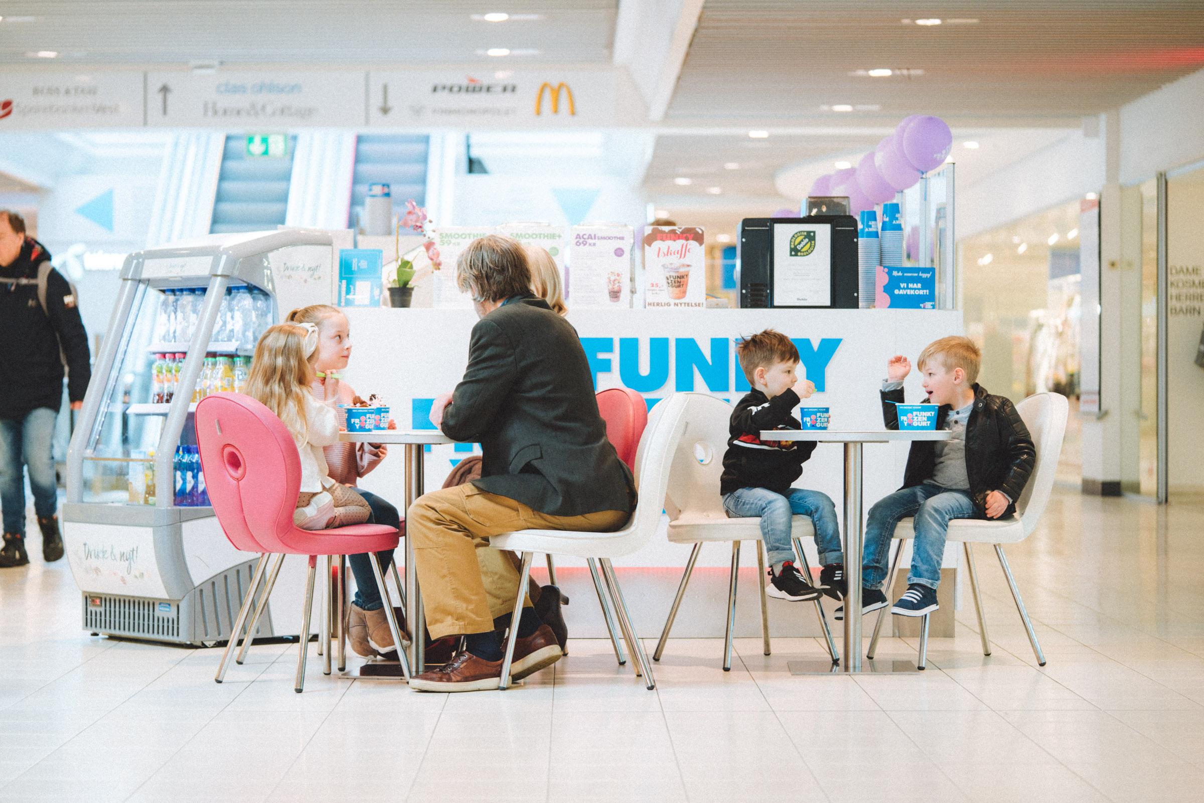 Familie som sitter ved et bord i en moderne kafé, med barn som spiser is og voksne som prater.