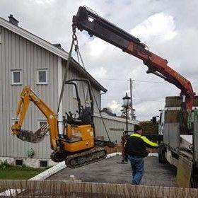 En gravemaskin heises opp av en kran mens en arbeider står ved siden av på en byggeplass.