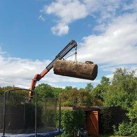 En kran løfter en stor tank over en trampoline i en hage med trær og en blå himmel.