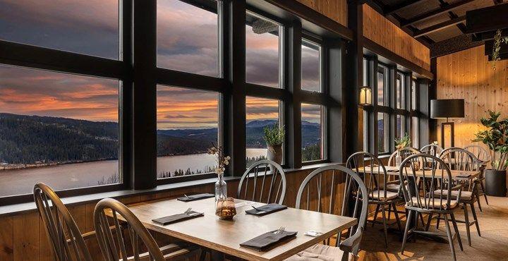 Koselig restaurant med store vinduer som gir utsikt over solnedgang og fjell. Bord og stoler i tre.