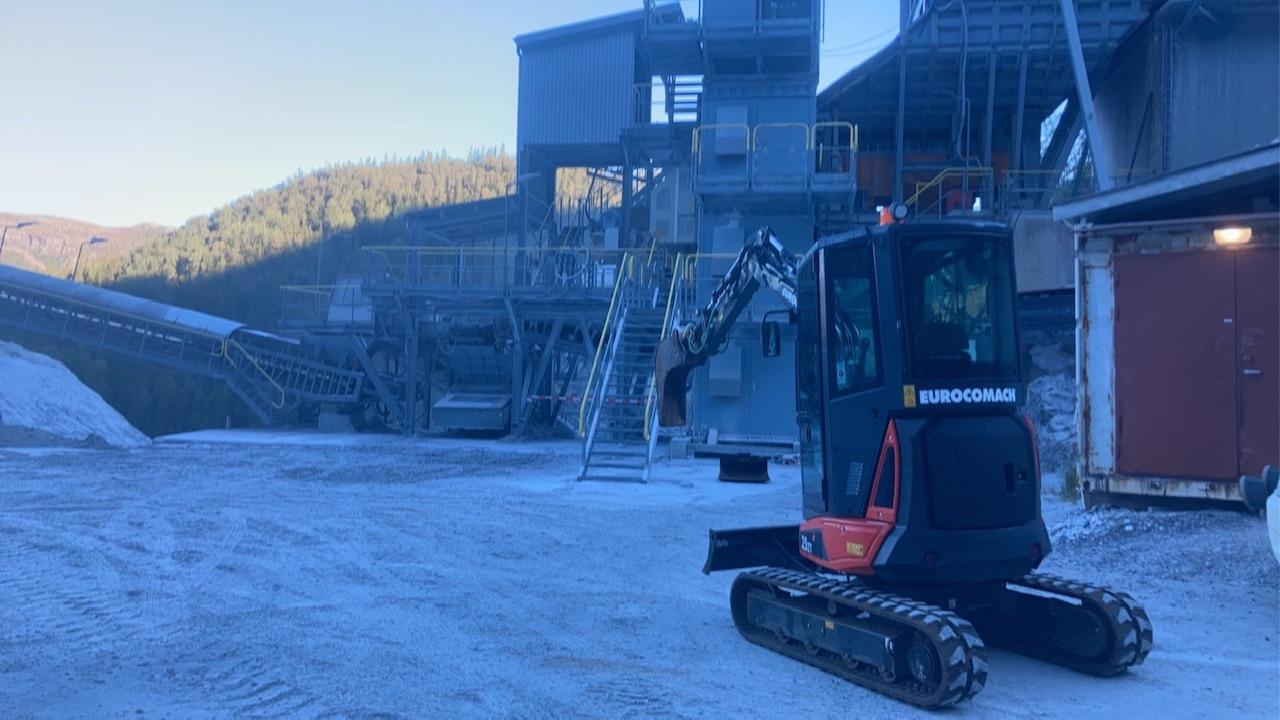 Gravemaskin på byggeplass med transportbånd og industrielt anlegg i bakgrunnen, omringet av fjell.