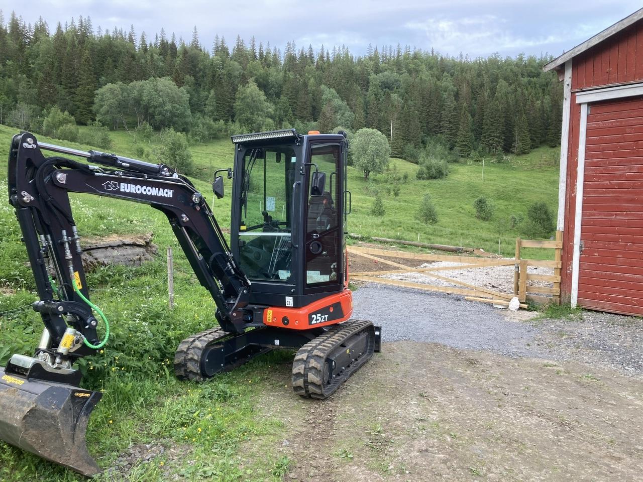 Liten gravemaskin parkert ved en rød låve, med grønt landskap og trær i bakgrunnen.