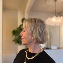 Kvinne med kort blond hår og gullsmykker, står i profil i et lyst rom med planter og lamper.