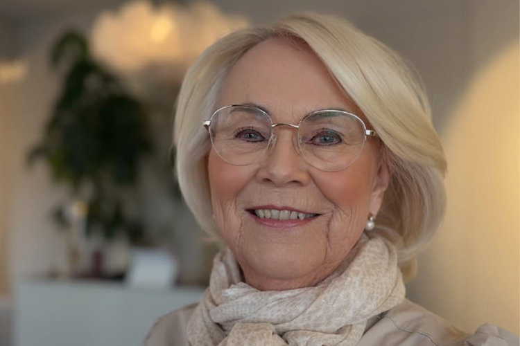 En smilende eldre kvinne med briller og lys hårfrisyre, iført en lys skjerf, står i et rom med planter.