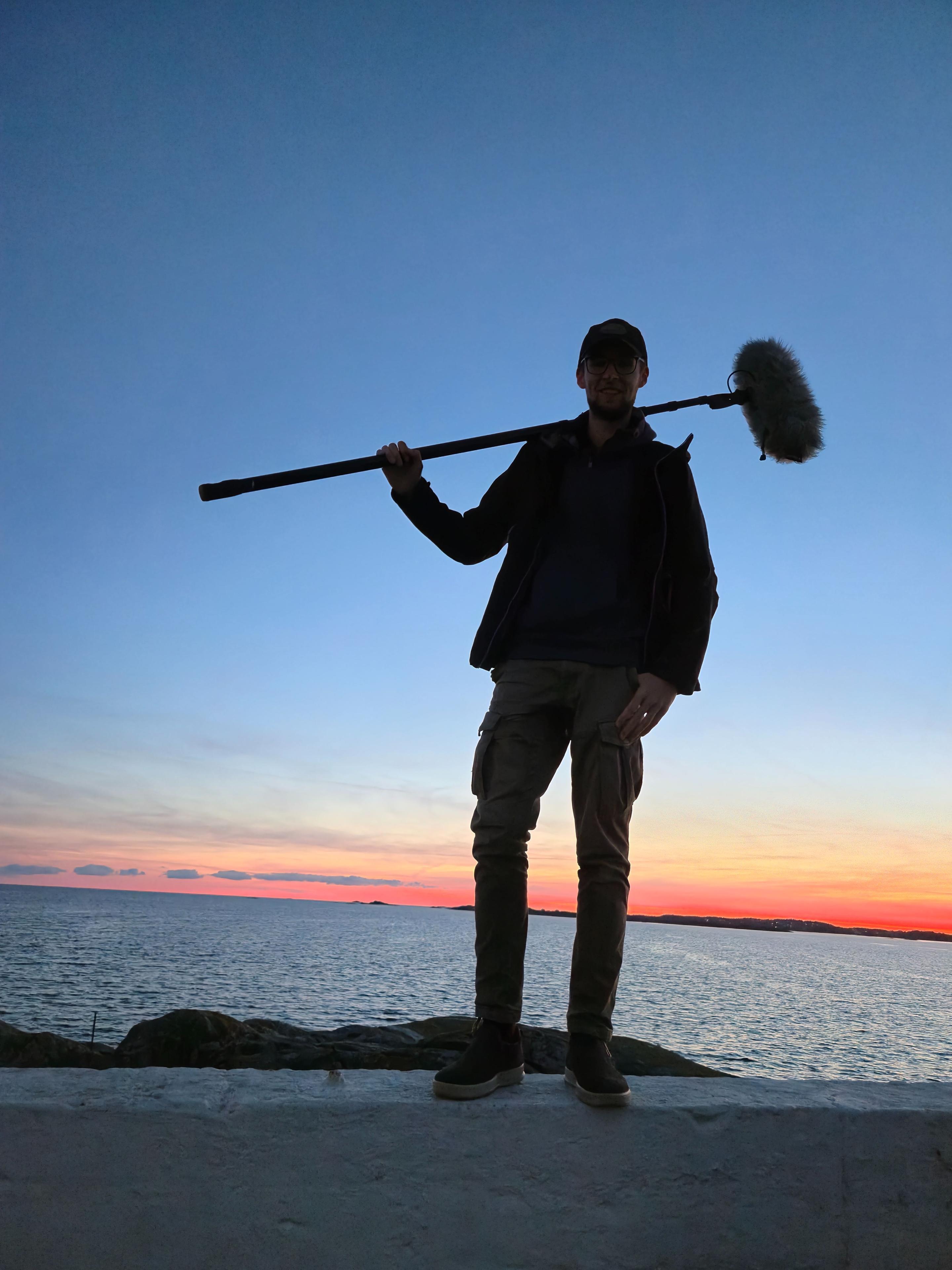 En mann står på en stein ved havet med en mikrofon på en stang, mot en vakker solnedgang.