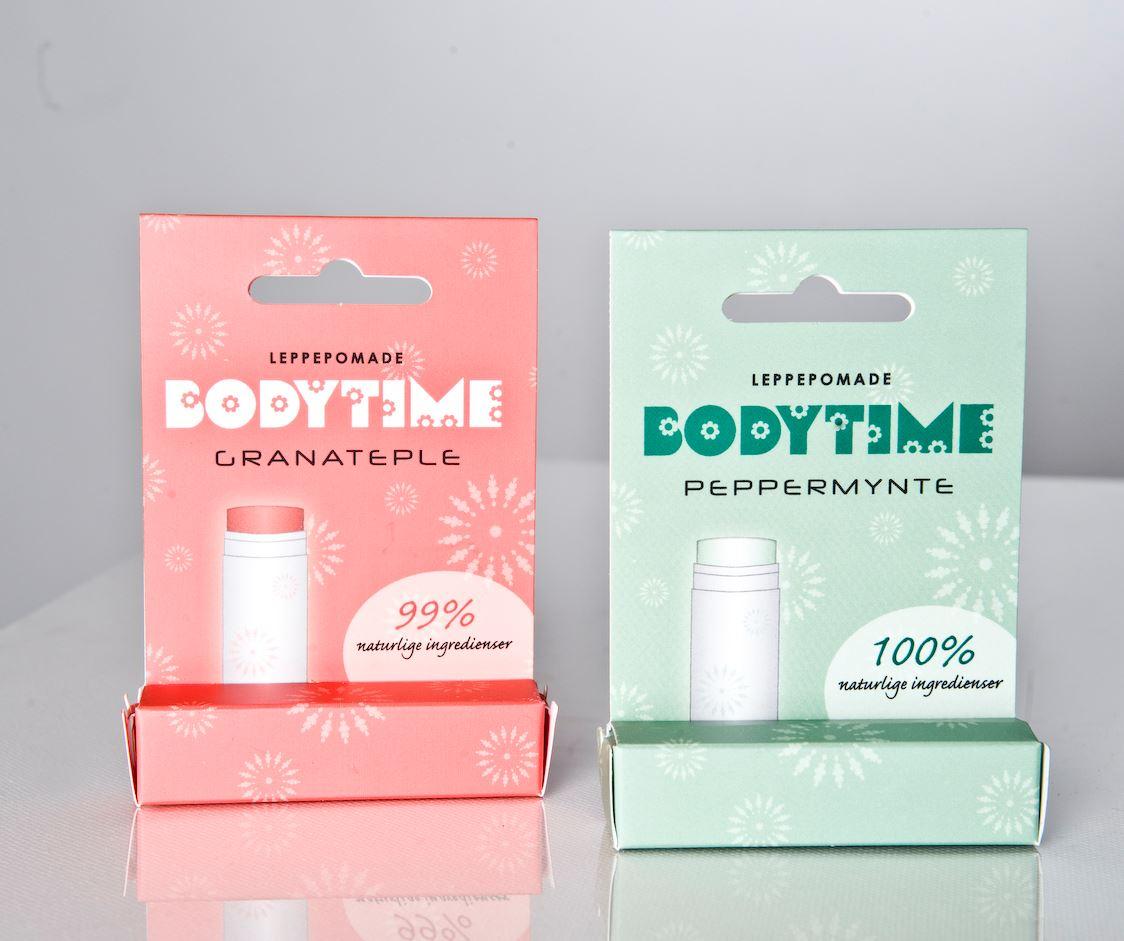 To pakker med leppepomade fra Bodytime, en med granateple og en med peppermynte, på en lys bakgrunn.