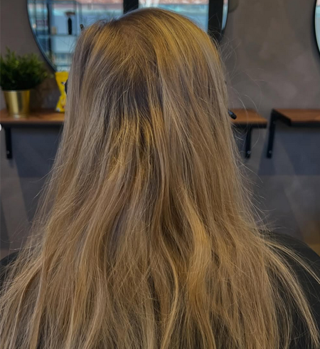 En kvinne med langt, lyst hår sett bakfra i en frisørsalong med speil og planter.