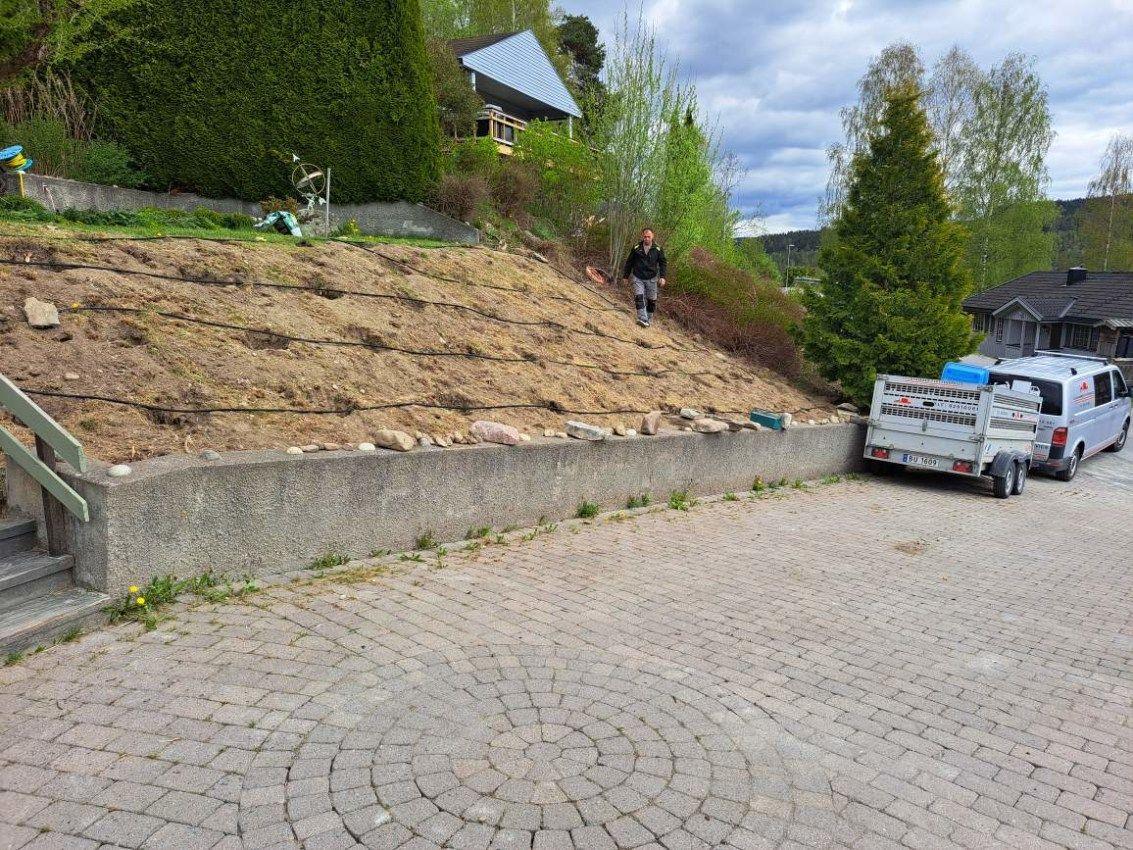 En skråning med nypløyd jord, en person som går nedover, og en bil med tilhenger parkert på brosteinsbelagt område.