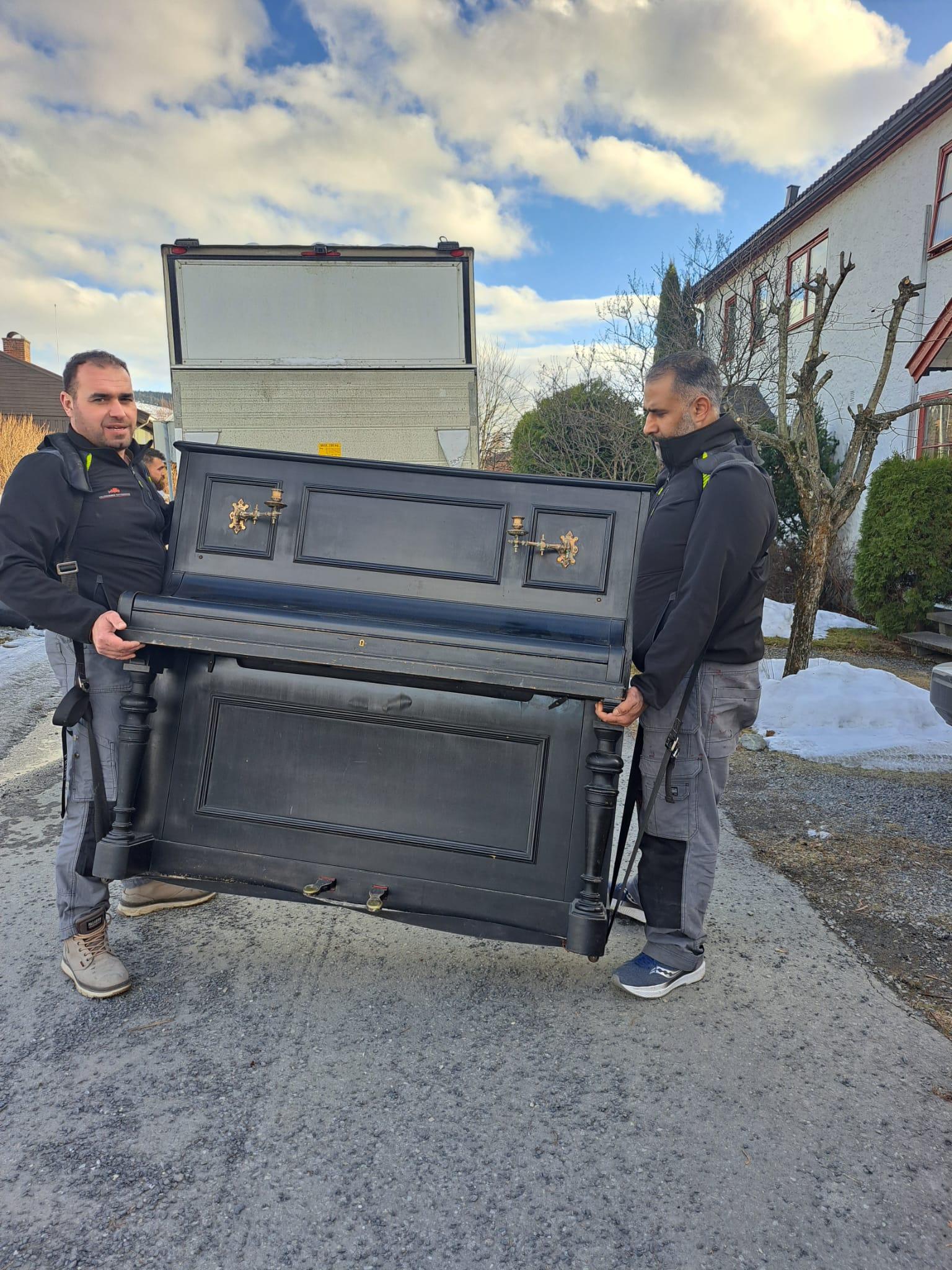 To menn bærer et svart piano ut av en lastebil på en snødekt vei. Himmelen er delvis skyet.