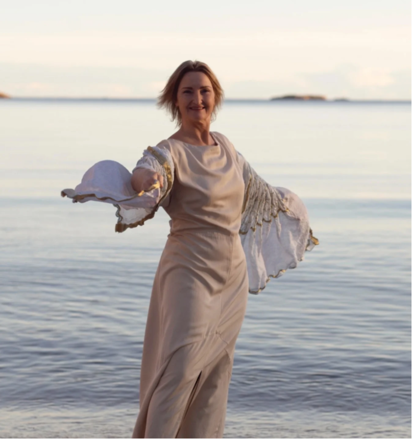 Kvinne i en elegant kjole med vide ermer står ved vannkanten med havet i bakgrunnen.