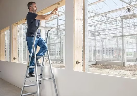 En mann står på en stige og installerer vinduer i et drivhus med mange glassvinduer.