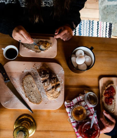 Frokostbord med brød, syltetøy, egg og kaffe, med hender som forbereder måltidet.