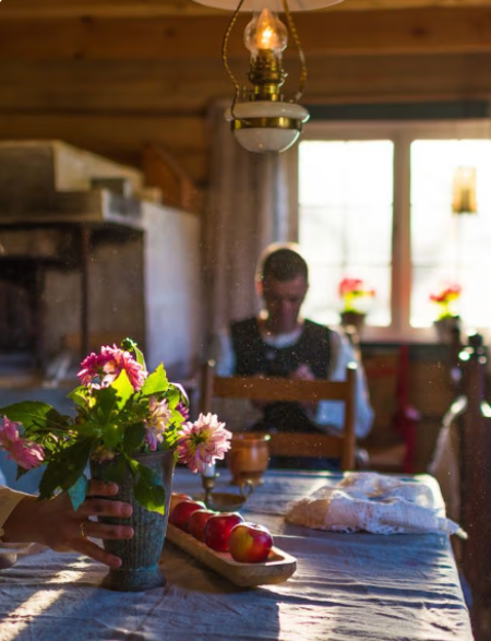 En koselig stue med en blomsterbukett på bordet, epler på en tallerken og en person som sitter i bakgrunnen.