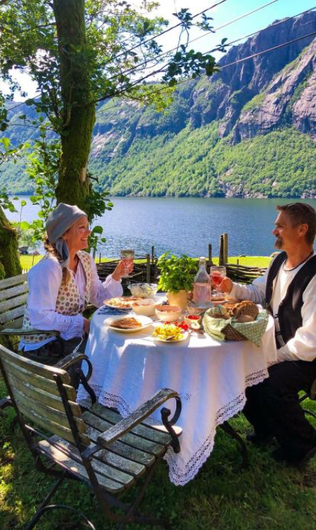 To personer i tradisjonelle klær sitter ved et bord med mat og drikke, med en vakker innsjø og fjell i bakgrunnen.