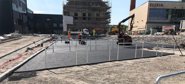 Byggeplass med arbeidere som legger asfalt og maskiner i bakgrunnen, samt en bygning under oppføring.