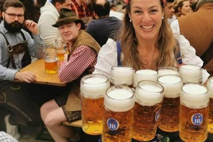 En kvinne i tradisjonell drakt holder flere store ølglass, mens menn i bakgrunnen smiler og sitter ved et bord.