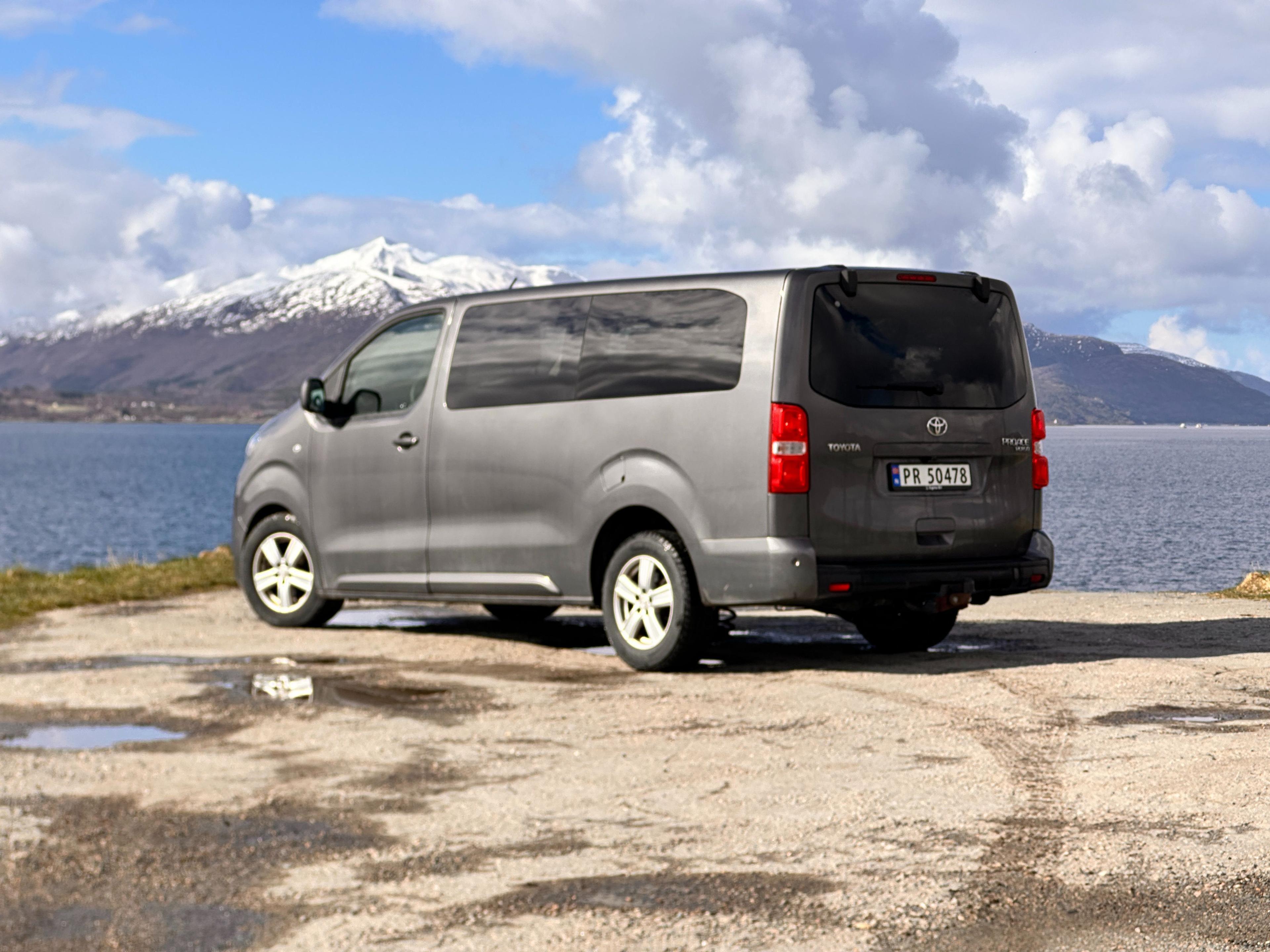 En grå varebil parkert ved vannkanten med snødekte fjell i bakgrunnen.