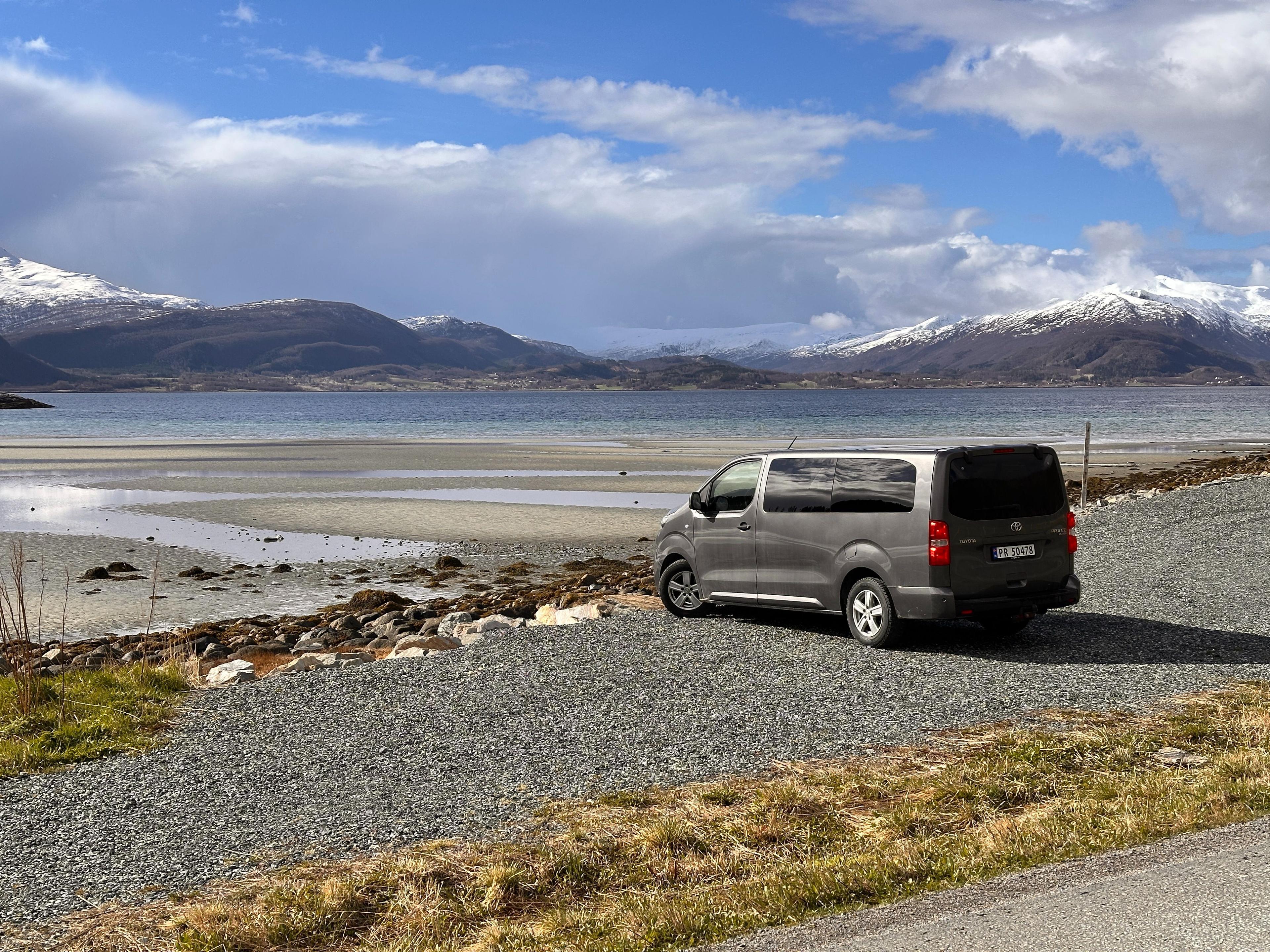 En grå varebil parkert ved en strand med fjell i bakgrunnen og blå himmel.