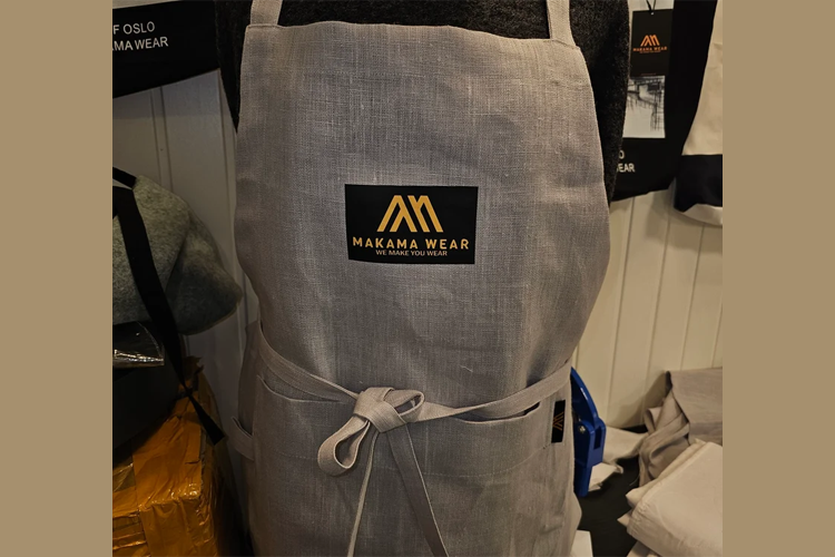 Grå forkle med Makana Wear-logo, plassert på en mannekeng i en butikk.