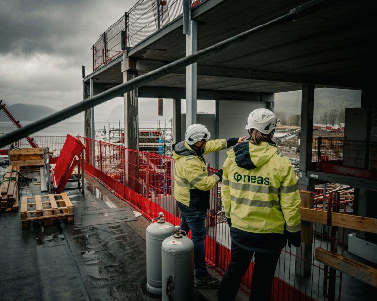 To personer i sikkerhetsutstyr på en byggeplass, med utsikt over vannet og byggearbeid i bakgrunnen.