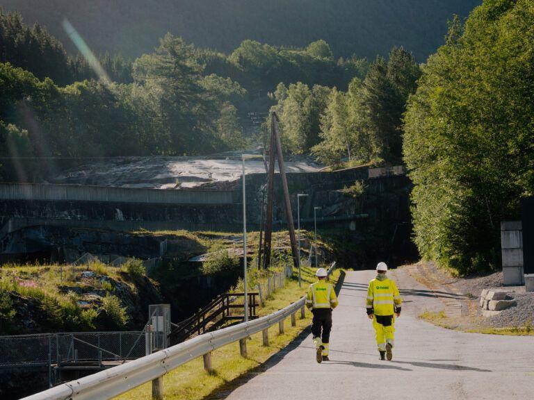 To personer i gule arbeidsklær går langs en vei med grønt landskap og en demning i bakgrunnen.