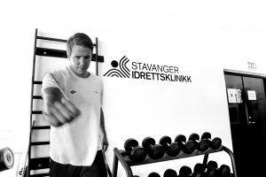 Mann som trener med vekter i Stavanger Idrettsklinikk, med treningsutstyr i bakgrunnen.