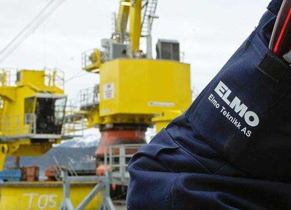 Arbeider i arbeidsklær med Elmo Teknikk AS-logo, med en offshore plattform i bakgrunnen.