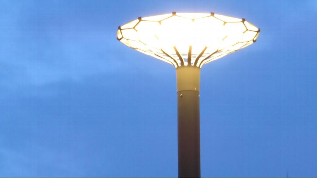 En moderne lyktestolpe med et blomsterlignende design som lyser opp en kveldshimmel.