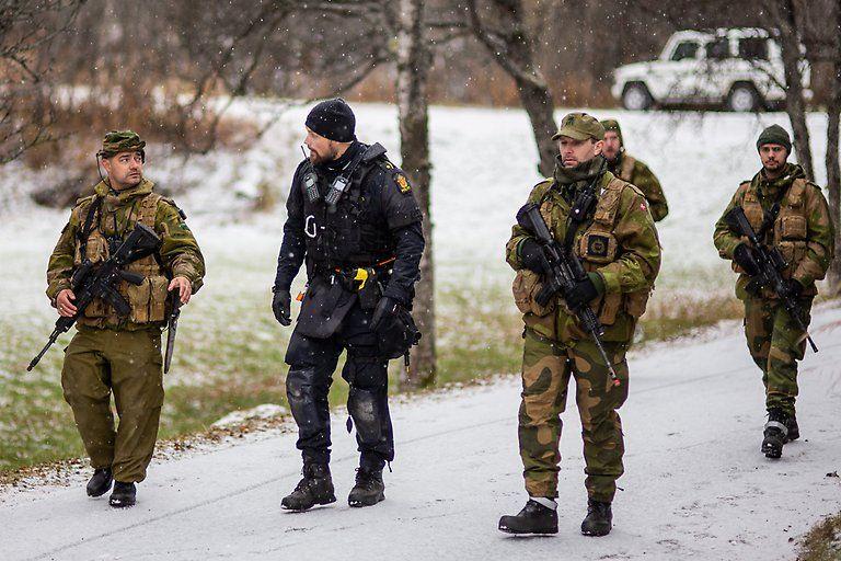 Fire soldater i militæruniformer og en politimann går langs en vei i snøvær. Trær og en bil er synlige i bakgrunnen.