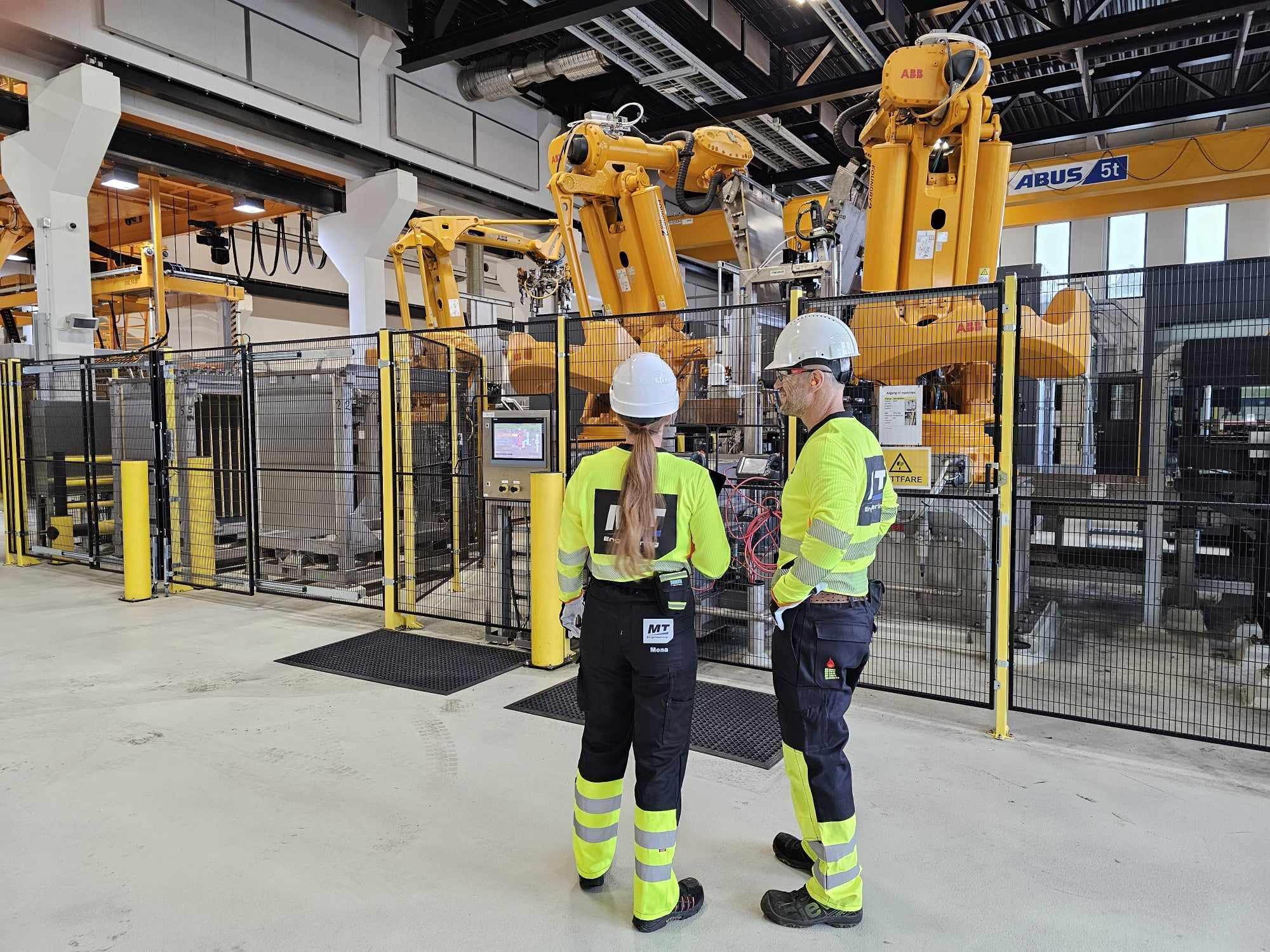 To personer i verneutstyr diskuterer ved en industriell robotcelle med gule roboter i bakgrunnen.