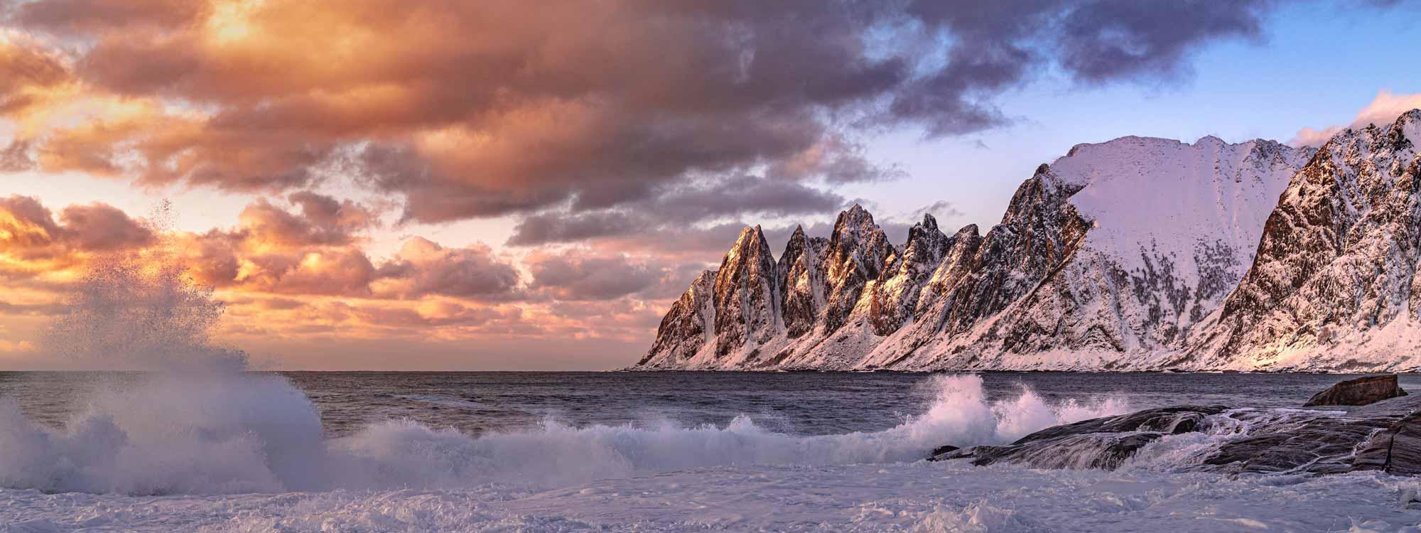 Vinterlandskap med snødekte fjell og bølger som slår mot kysten under en fargerik solnedgang.