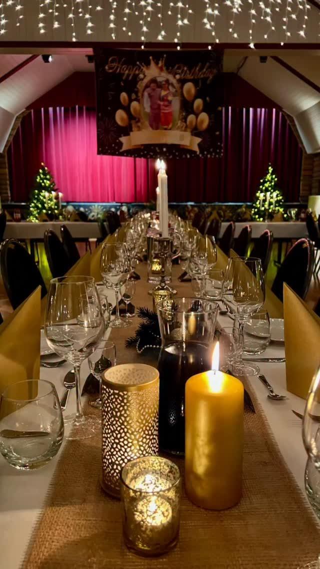 Et festdekket bord med stearinlys, glass og dekorasjoner, i en festlokale med julepynt og lys.
