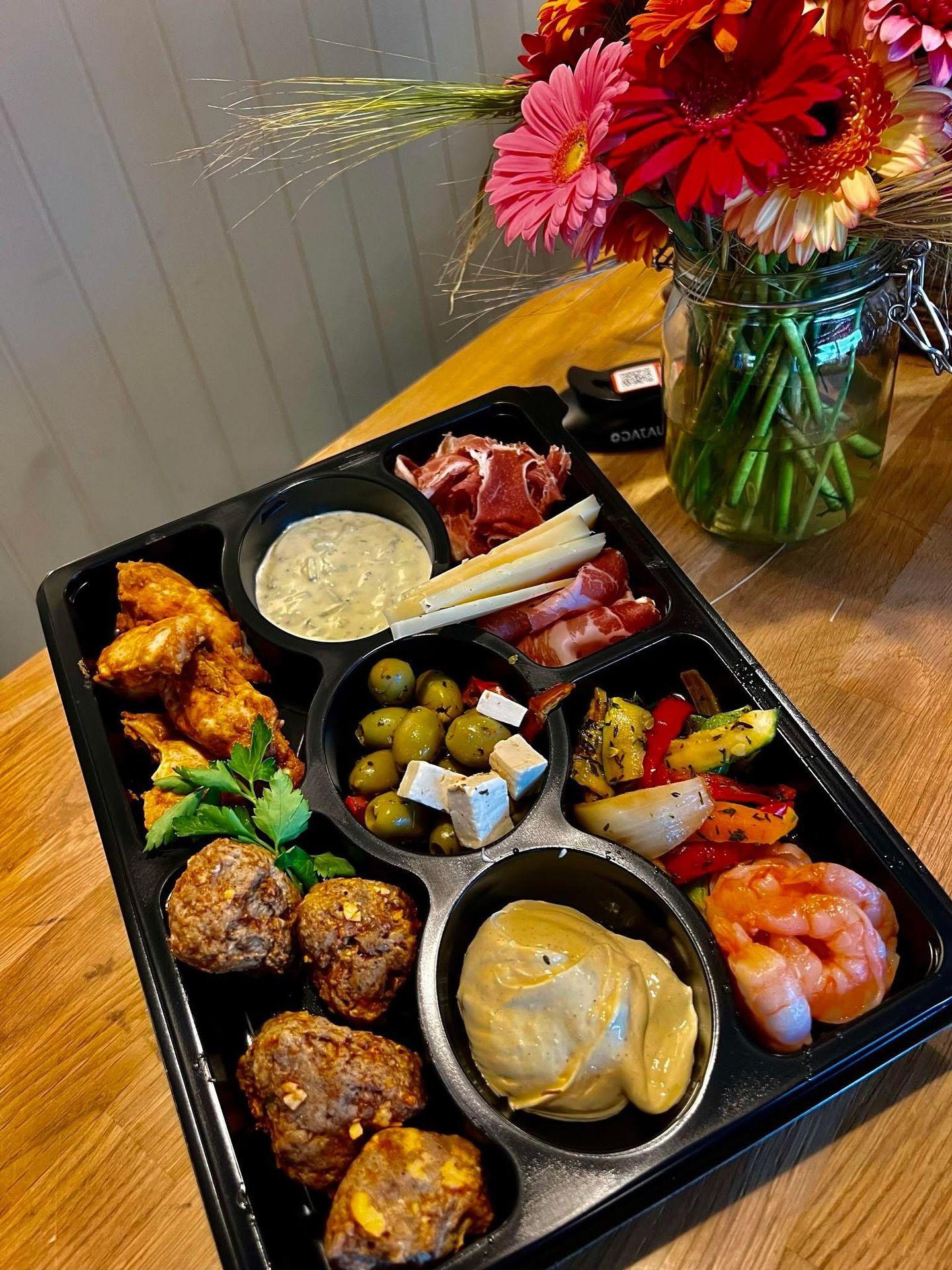 Et brett med variert tapas, inkludert kjøttboller, reker, oliven, grønnsaker og dipp, med blomster i bakgrunnen.