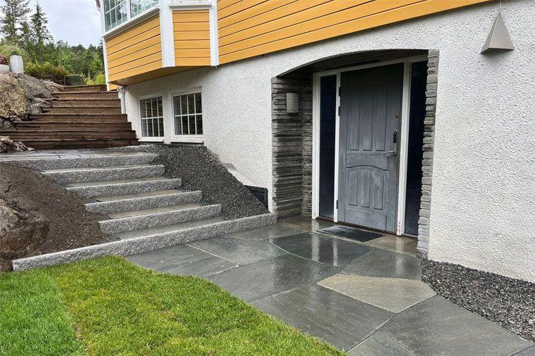 Inngangspartiet til et hus med grå steintrapper, en tredør og en plen i forgrunnen.