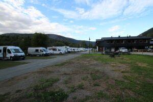Campingplass med flere campingvogner og en bygning i bakgrunnen, omringet av fjell og blå himmel.