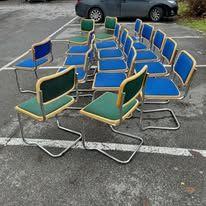 Flere stoler med blå og grønne seter står i en parkeringsplass med biler i bakgrunnen.