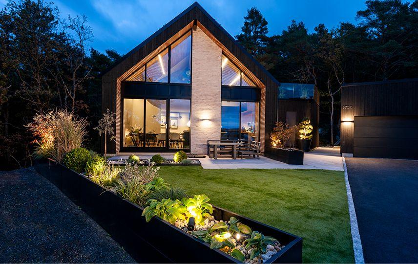 Moderne hus med store vinduer, beliggende i en hage med grønt gress og belysning om kvelden.
