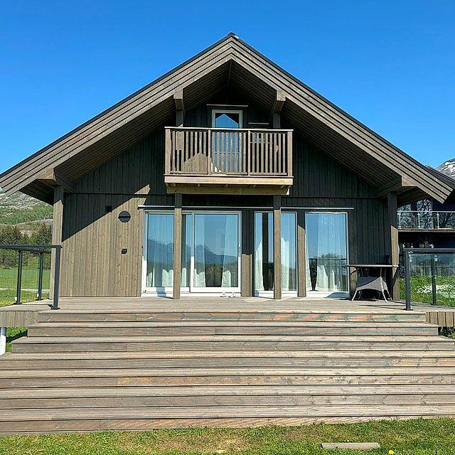 Moderne hytte med balkong og store vinduer, omgitt av grønt landskap og fjell i bakgrunnen.