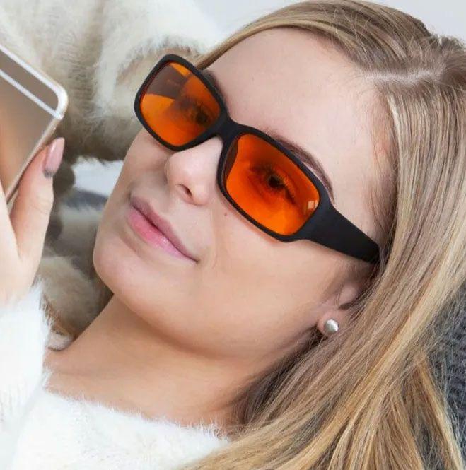 Ung kvinne med oransje solbriller som ser på telefonen sin. Hun har langt, lyst hår og en hvit genser.