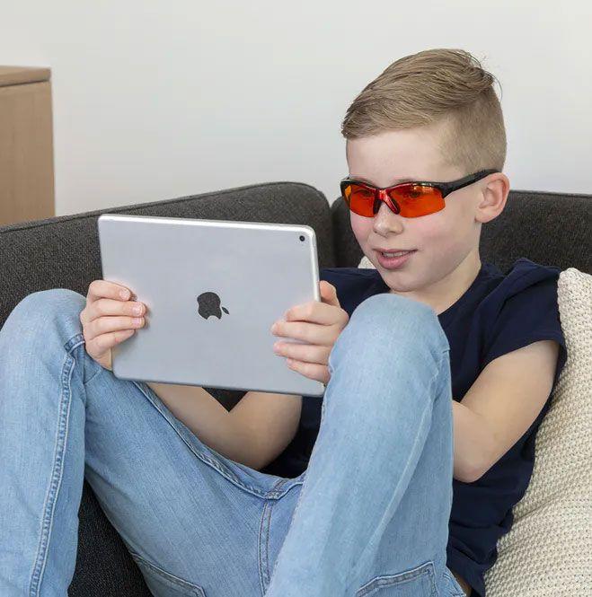 En gutt med solbriller sitter på en sofa og bruker en iPad.