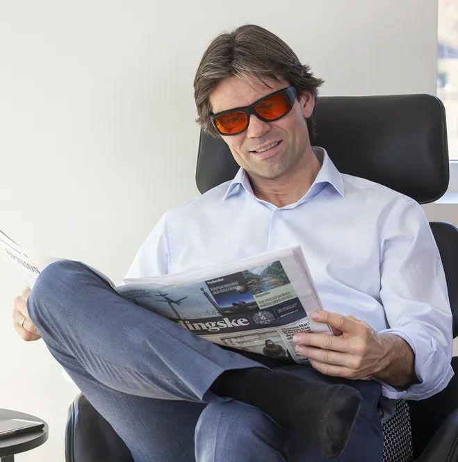 Mann med solbriller som leser avis mens han sitter komfortabelt i en stol.