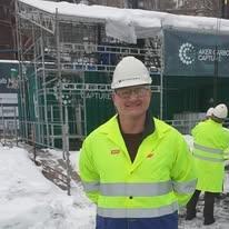 En smilende mann i gul arbeidsjakke og hjelm står foran en byggeplass dekket av snø.